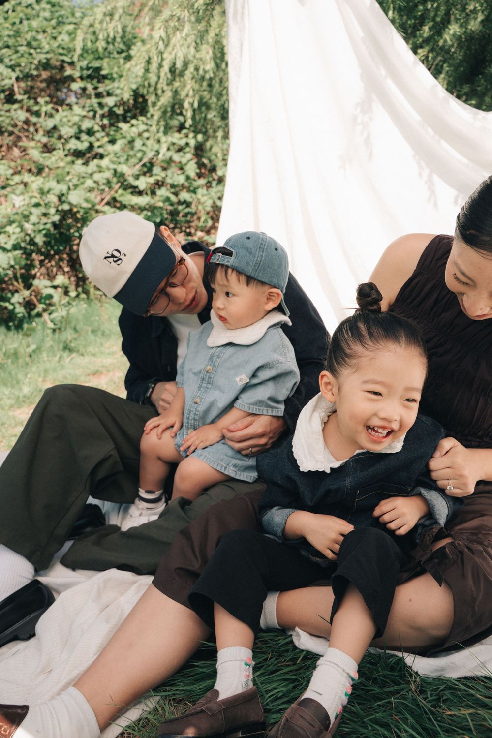 Happy family with two young children sitting outdoors near a white canopy tent surrounded by lush greenery.