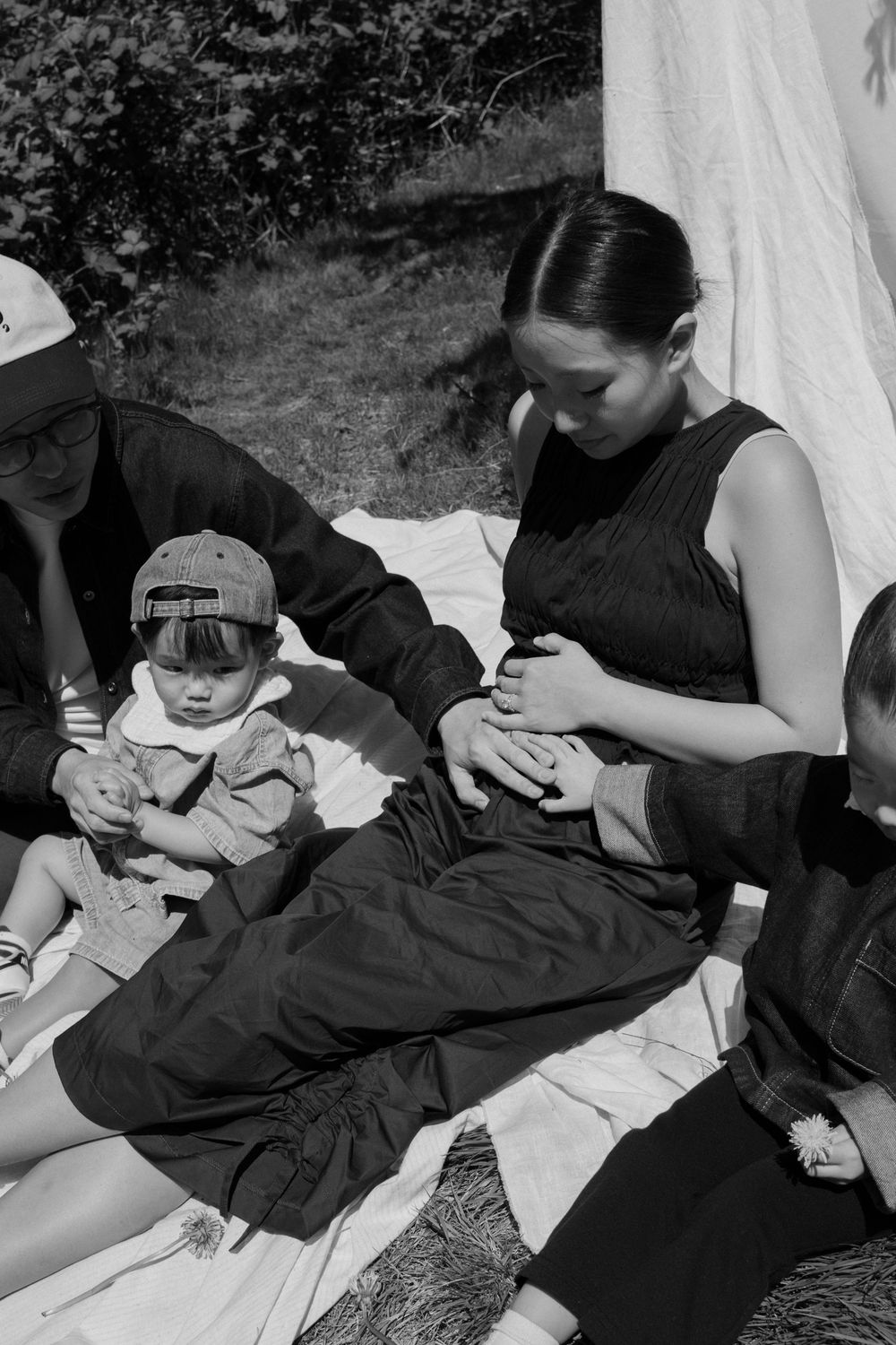 Black and white photo of a family sitting outdoors on a blanket near a tent, with a baby wearing a cap.