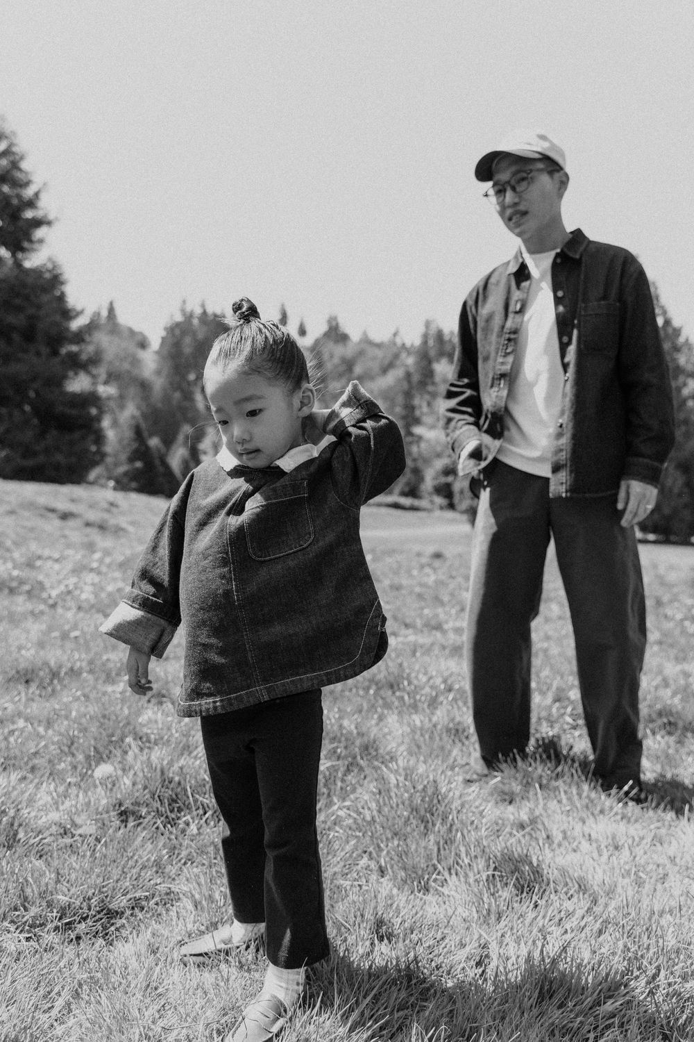Black and white photo of a young Asian child in a denim jacket playing outdoors while an adult man watches nearby.