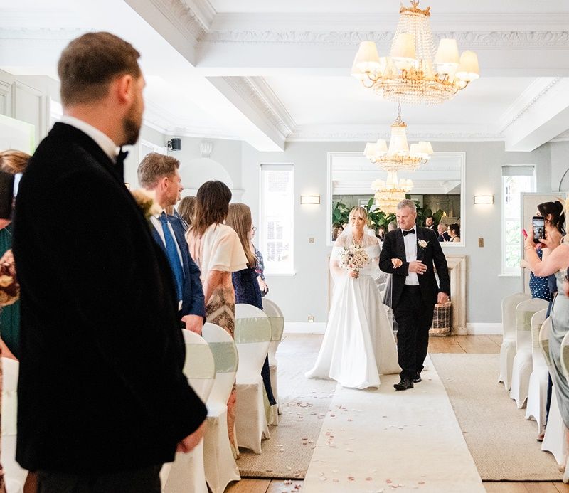 Bride walks down the aisle with her father in an elegant chandelier-lit wedding venue.