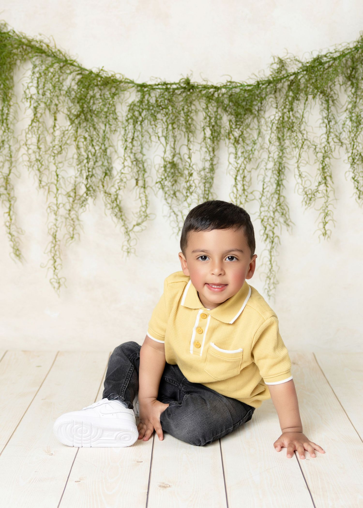 Young boy in yellow polo shirt and dark jeans sits smiling on white wood floor with green garland backdrop.