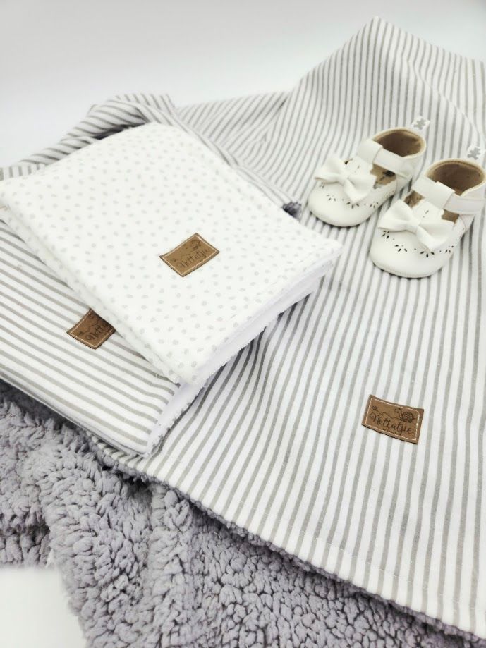 Gray striped baby blanket set with white dotted swaddle and white bow baby shoes on soft textured surface.