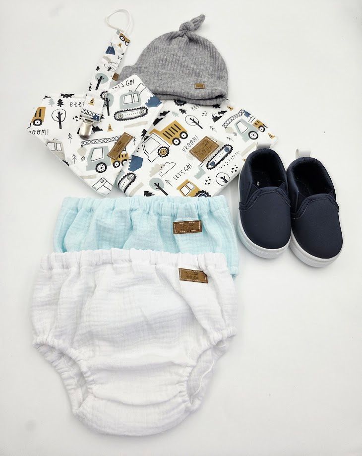 Baby boy outfit flat lay with printed bib, gray knot hat, muslin bloomers, and navy slip-on shoes.
