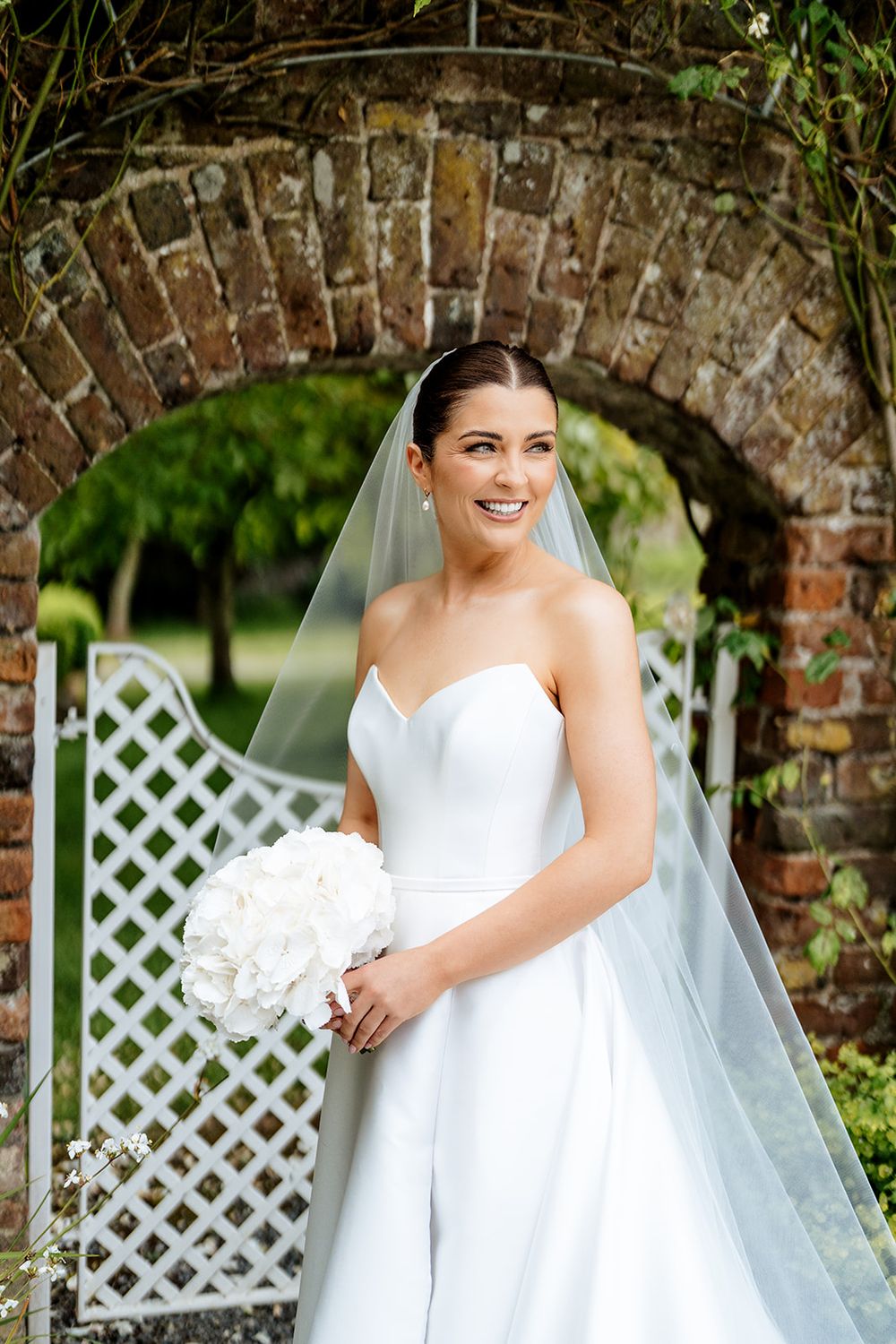 A bride in a white wedding dress stands by a white lattice garden trellis against a stone wall backdrop.