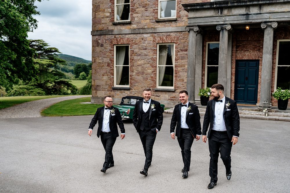 Groomsmen walk and pose together outside a rustic brick building in formal black suits during a wedding photoshoot.