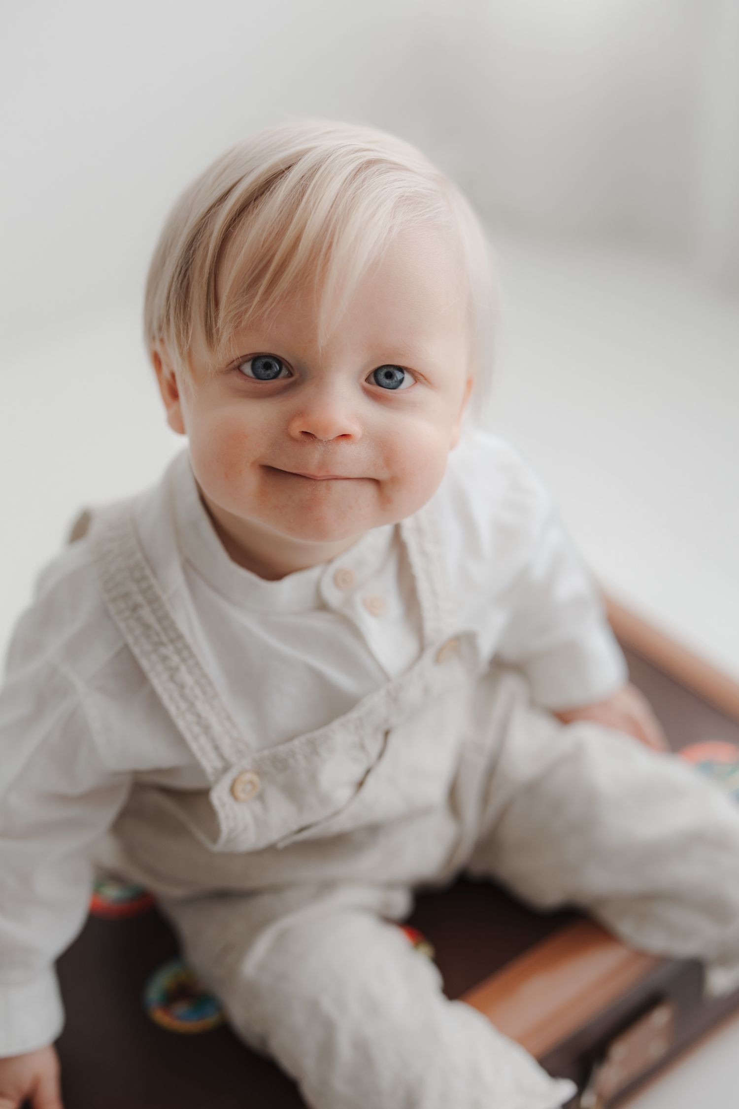 Smiling baby with blue eyes and blonde hair wearing a white linen outfit, sitting on the floor.