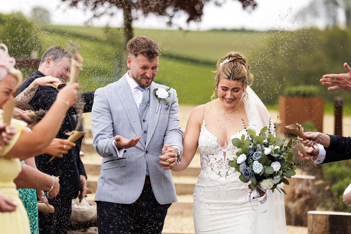 Bride and groom walking together at outdoor wedding ceremony while guests throw confetti in celebration.