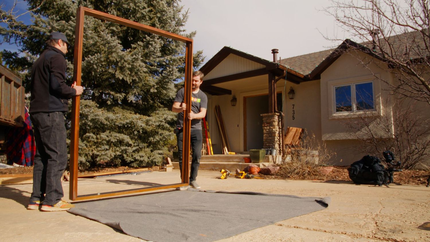 A person stands next to a wooden frame structure in front of a tan house with blue windows on a sunny day.