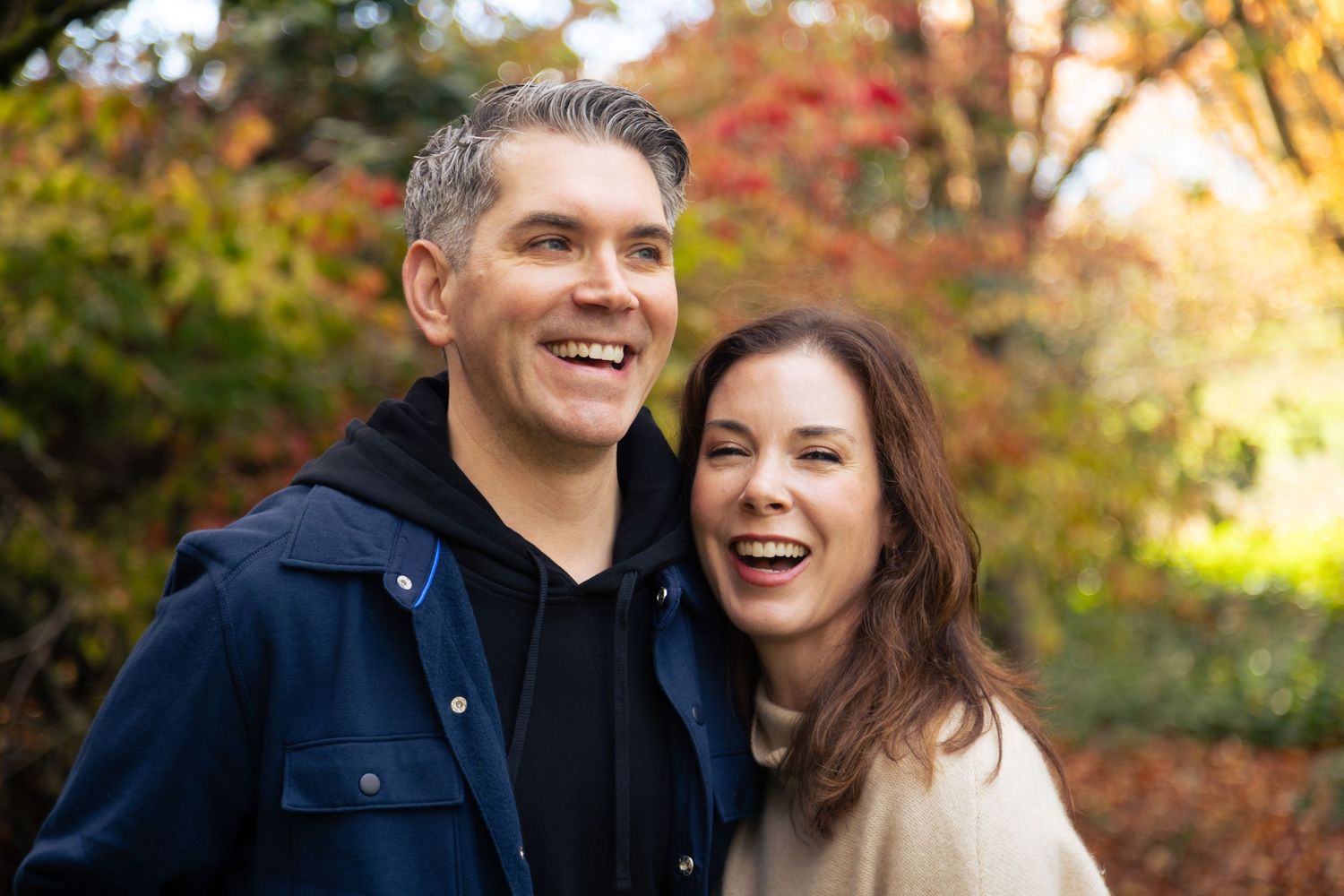 Smiling couple laughing together outdoors in autumn park with colorful fall foliage in background.