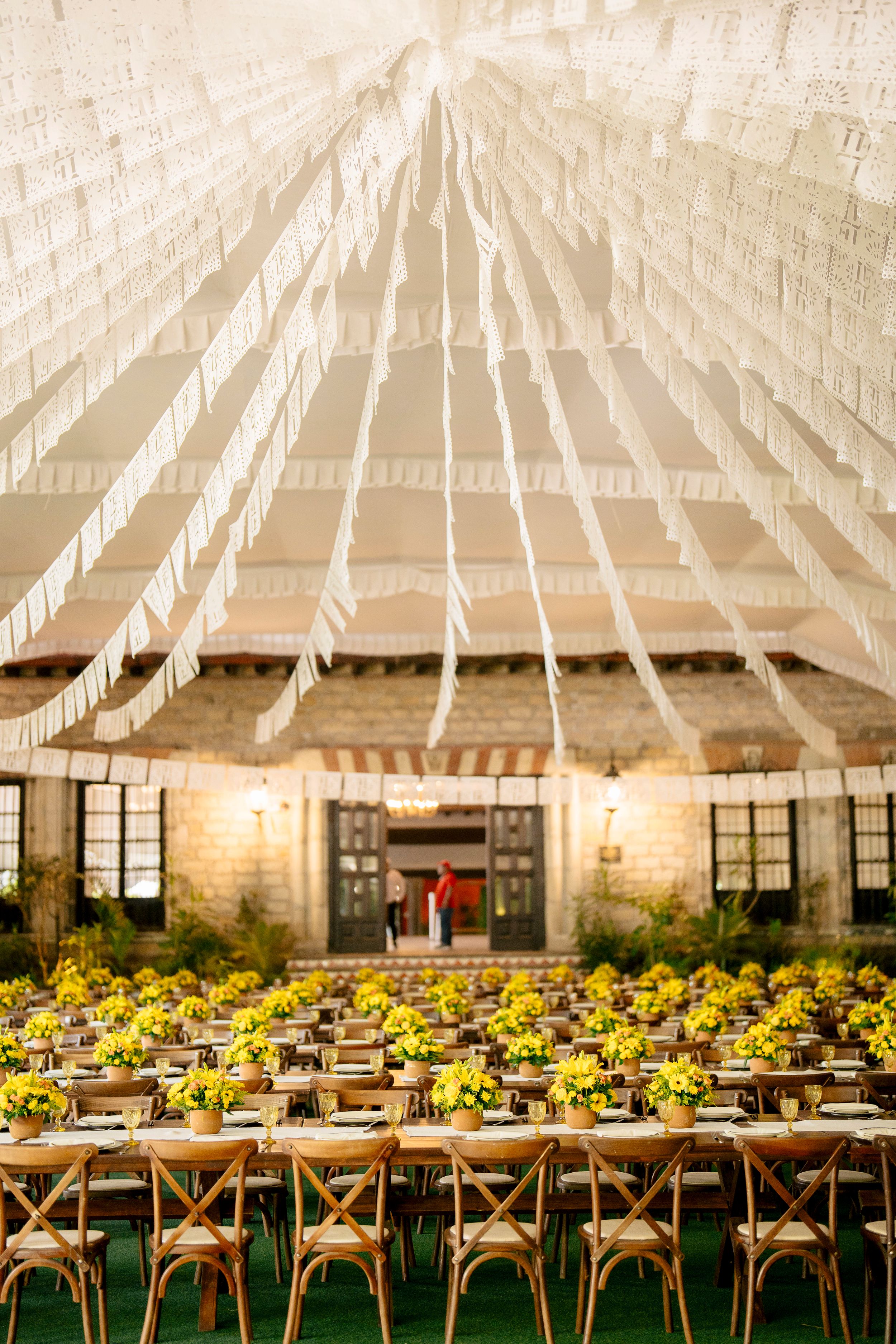 Elegant outdoor wedding reception tent with yellow floral centerpieces, wooden cross-back chairs, and white ribbon ceiling decor.