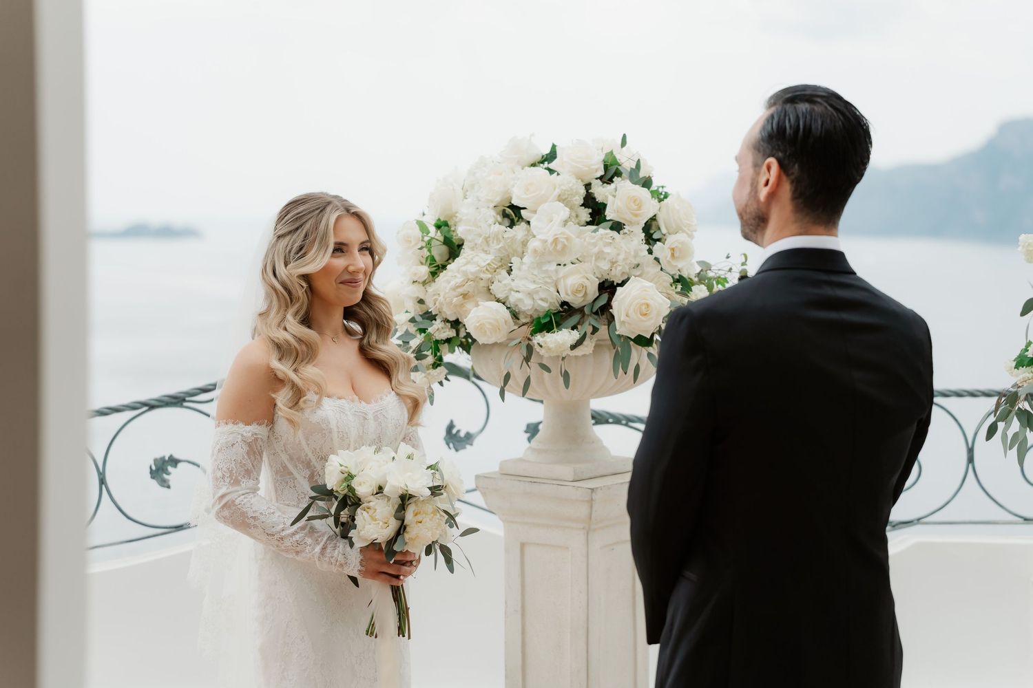 Bride in off-shoulder gown holds white bouquet facing groom in black suit at elegant outdoor wedding ceremony.
