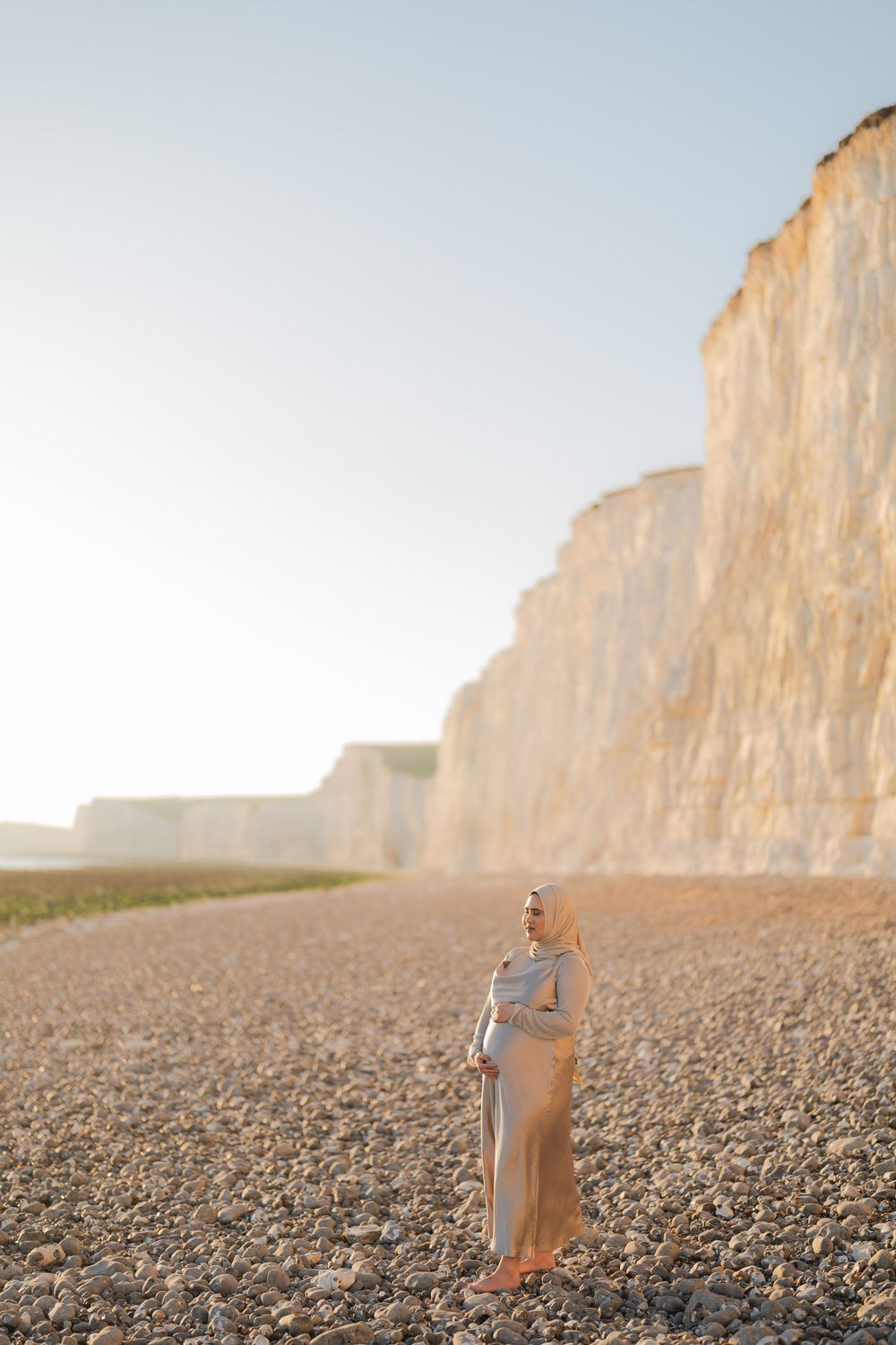 Pregnant woman in flowing dress stands on pebble beach beside towering white chalk cliffs at golden hour.