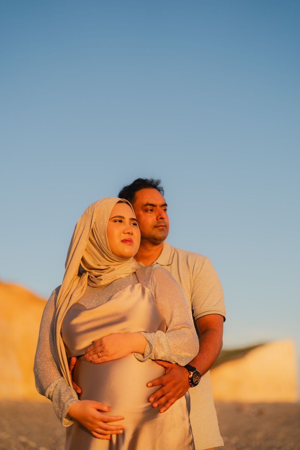 Pregnant Muslim couple embracing outdoors at golden hour, woman in hijab, man holding her baby bump against desert backdrop.
