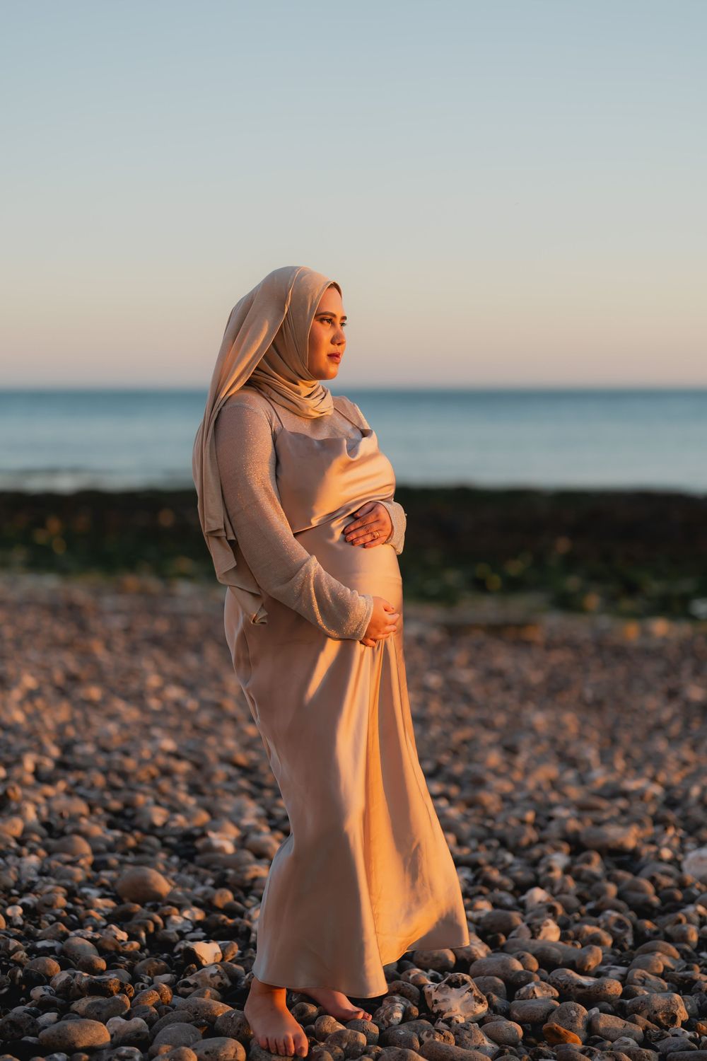 Pregnant Muslim woman in beige hijab and dress cradling her bump on a pebble beach at sunset.