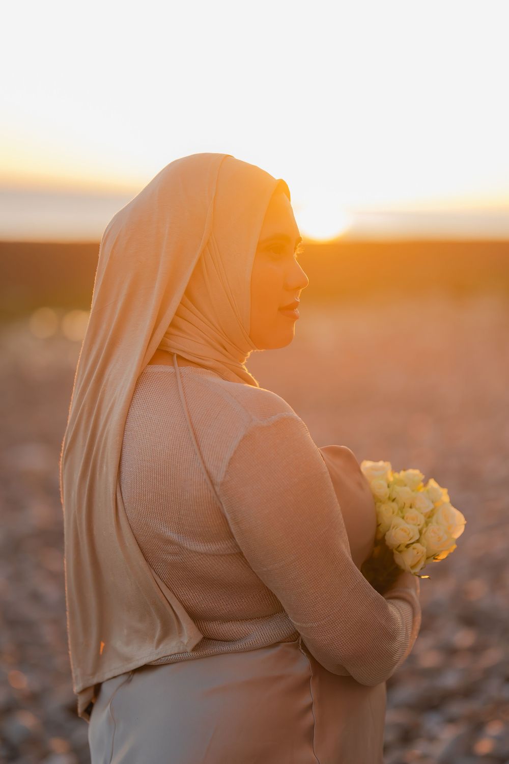 A woman in a beige hijab and dress holds yellow roses, standing in profile against a golden sunset backdrop.