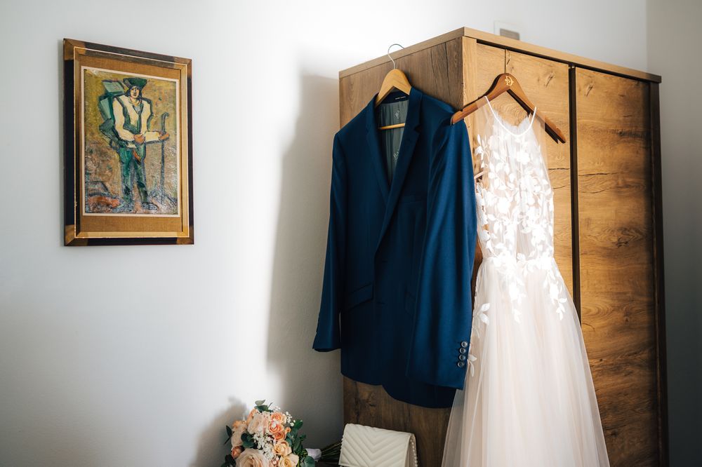 Blue groom suit jacket and white lace wedding dress hanging on wooden wardrobe before wedding ceremony.