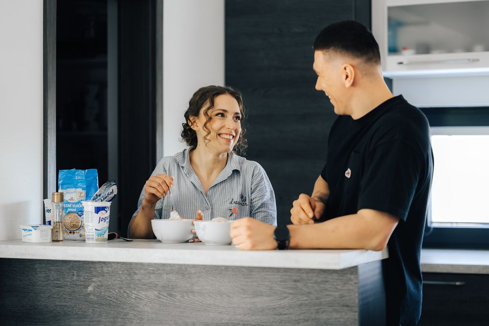 Two people smiling and chatting over bowls at a modern kitchen counter with dairy products nearby.