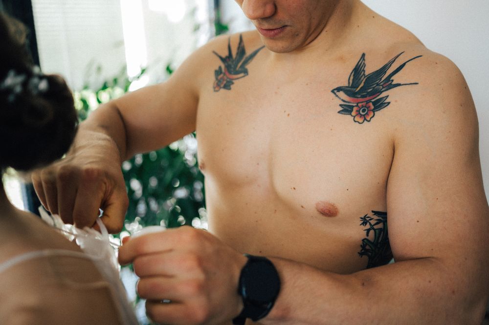 Shirtless man with bird tattoos on chest and arm fastening a white garment.
