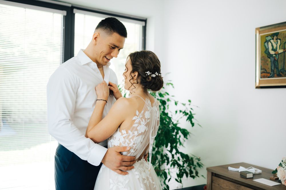 A couple in wedding attire share a tender moment, the bride in lace gown and groom in white shirt, embracing indoors.
