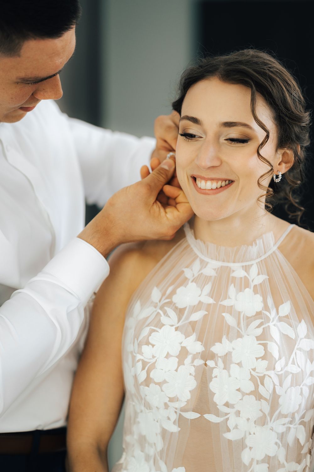 Groom helping smiling bride put on earring, bride wearing white floral lace halter wedding dress.