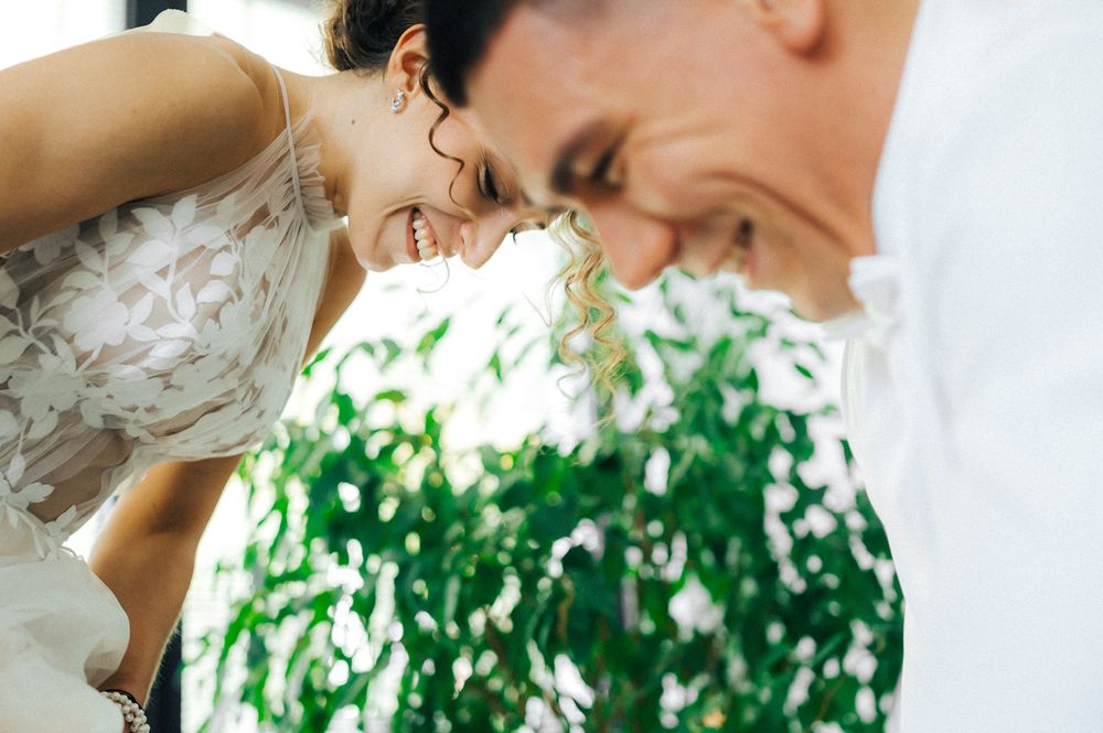 Smiling bride in lace wedding dress leaning over green bouquet with groom in white shirt.