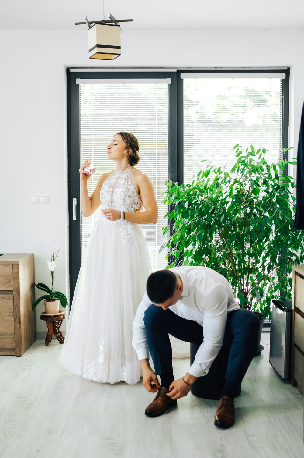 Bride in white gown holds glass while groom ties his brown dress shoes before wedding ceremony.