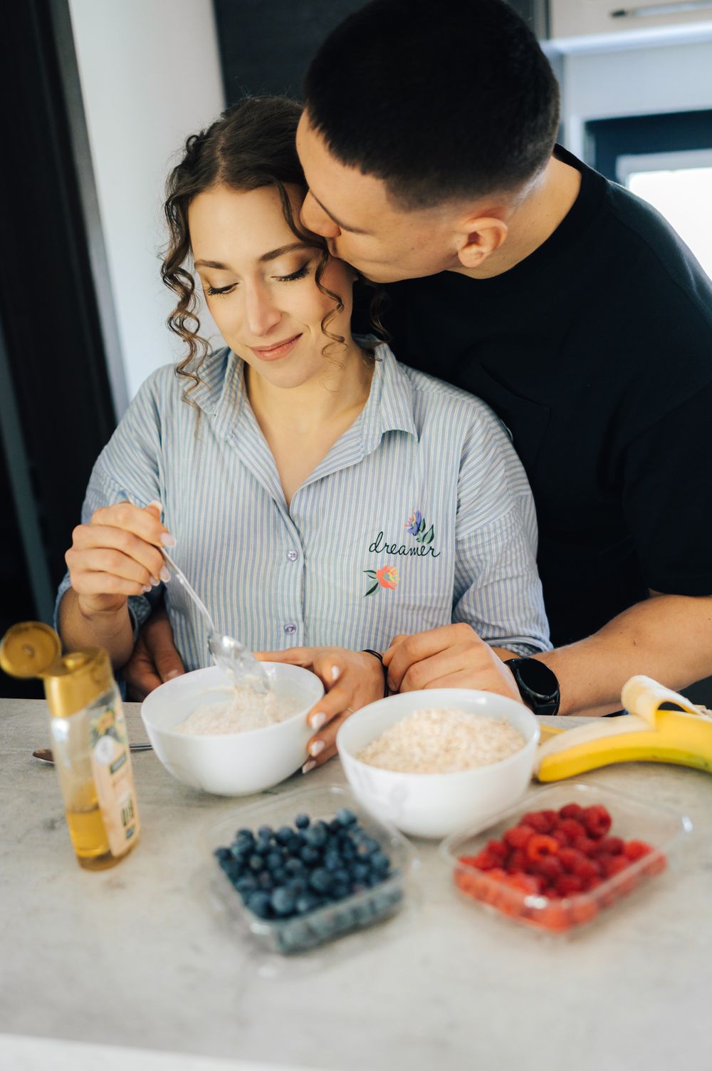 Couple sharing a tender moment at kitchen counter with bowls of oatmeal, blueberries, raspberries, and banana.