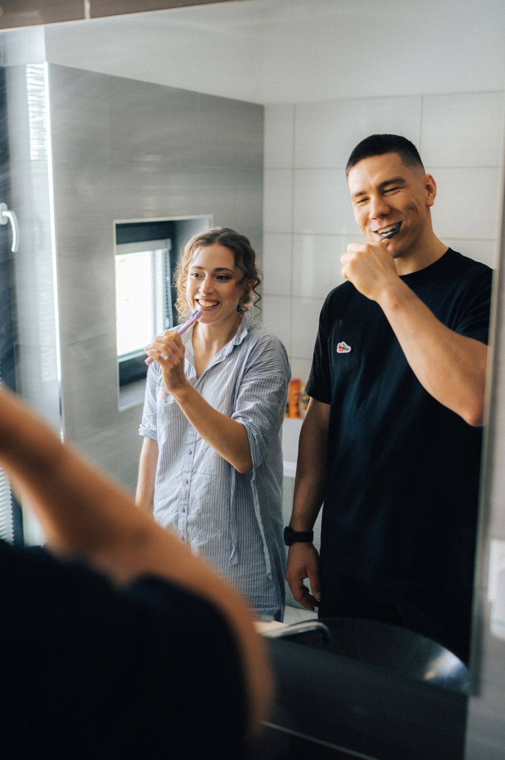 Couple brushing teeth together in modern bathroom mirror, smiling in pajamas and black t-shirt during morning routine.