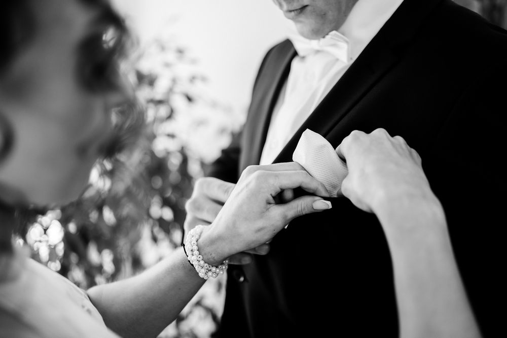 Black and white photo of a woman fastening a boutonniere on a groom's lapel at a wedding ceremony.