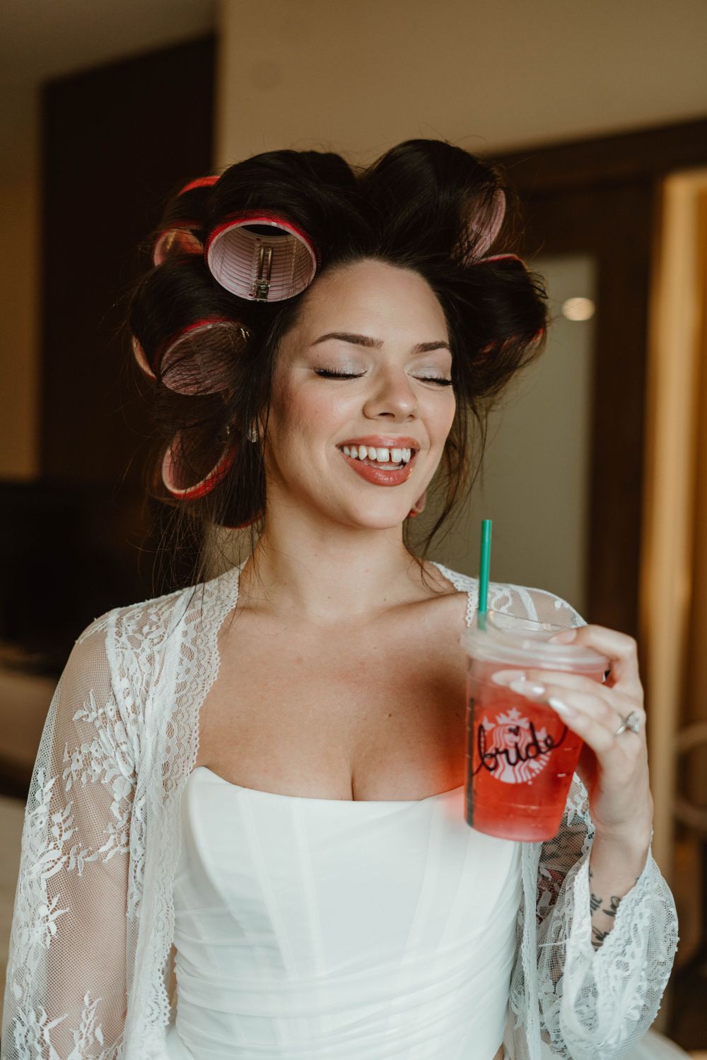 A joyful bride in hair rollers and white lace robe smiles while holding a pink Sonic drink during wedding preparations.