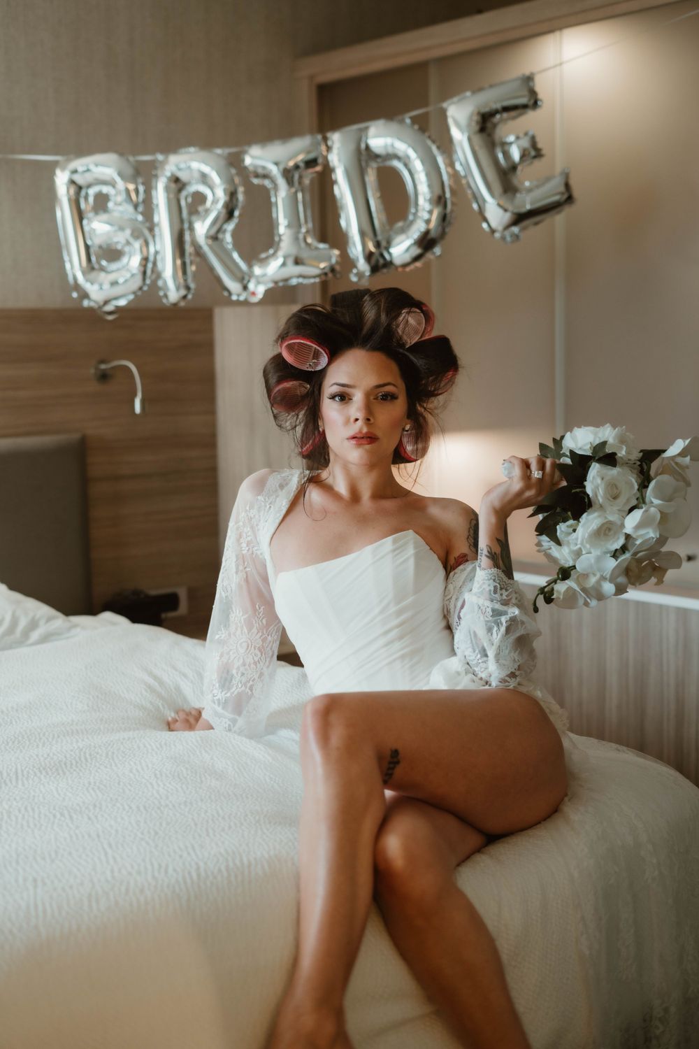 Boudoir bridal photo with silver BRIDE balloons, hair rollers, and elegant white lingerie in a hotel room setting.