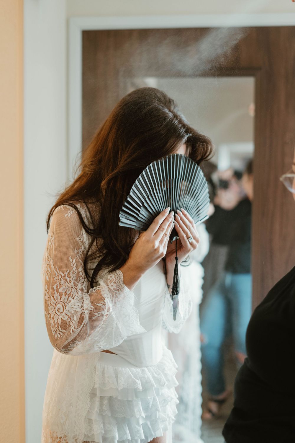 Person in white lace dress holding a decorative black folding fan in front of their face.