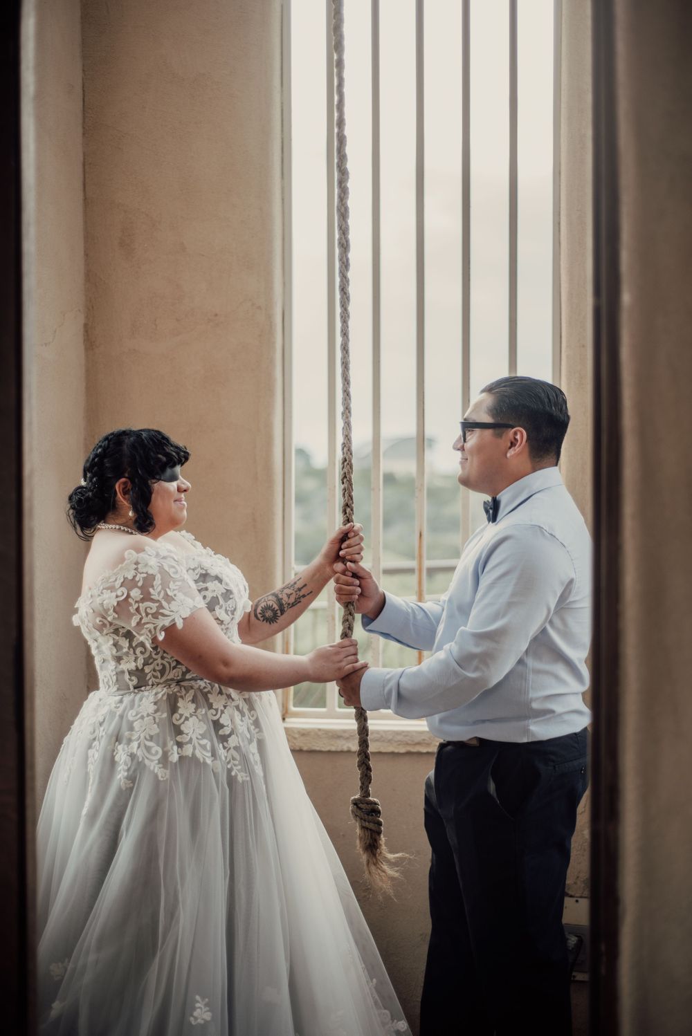 A couple shares a romantic moment in formal wedding attire holding an ornate sword near a columned window.