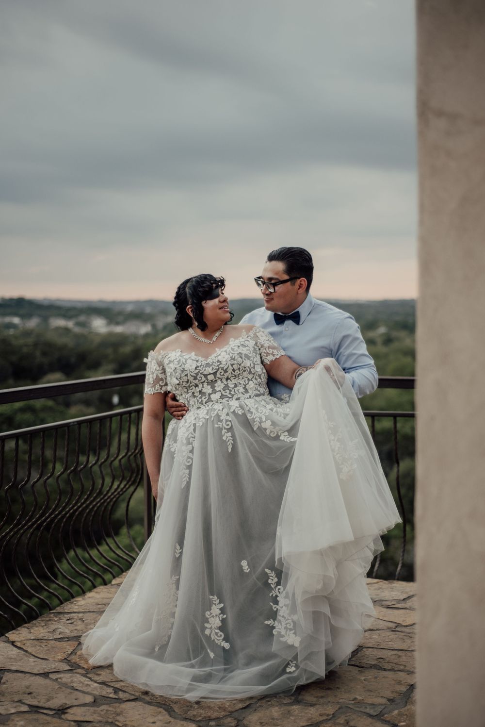 A couple shares a romantic moment on a balcony overlooking scenic hills, the bride wearing an elegant flowing lace gown.