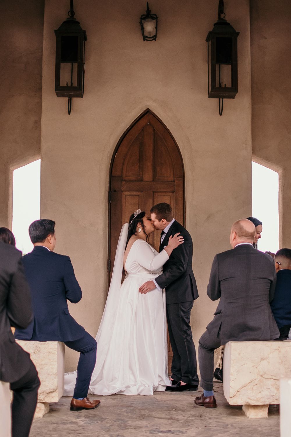 A romantic wedding ceremony takes place in an archway with lanterns as guests watch from stone benches.