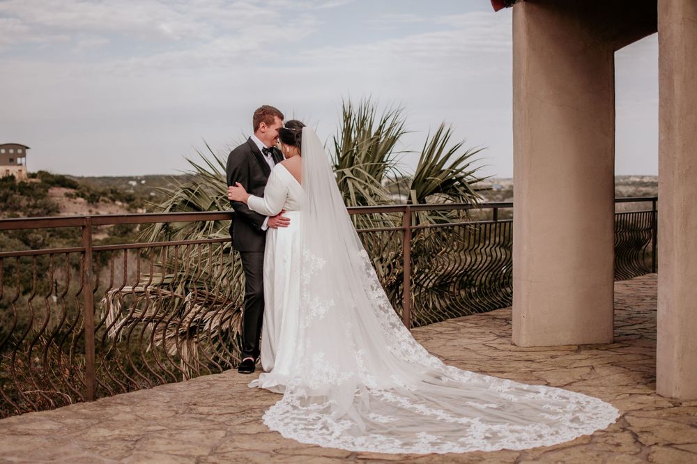 A romantic wedding couple embraces on a balcony overlooking coastal landscapes with palm trees and an elegant lace train.
