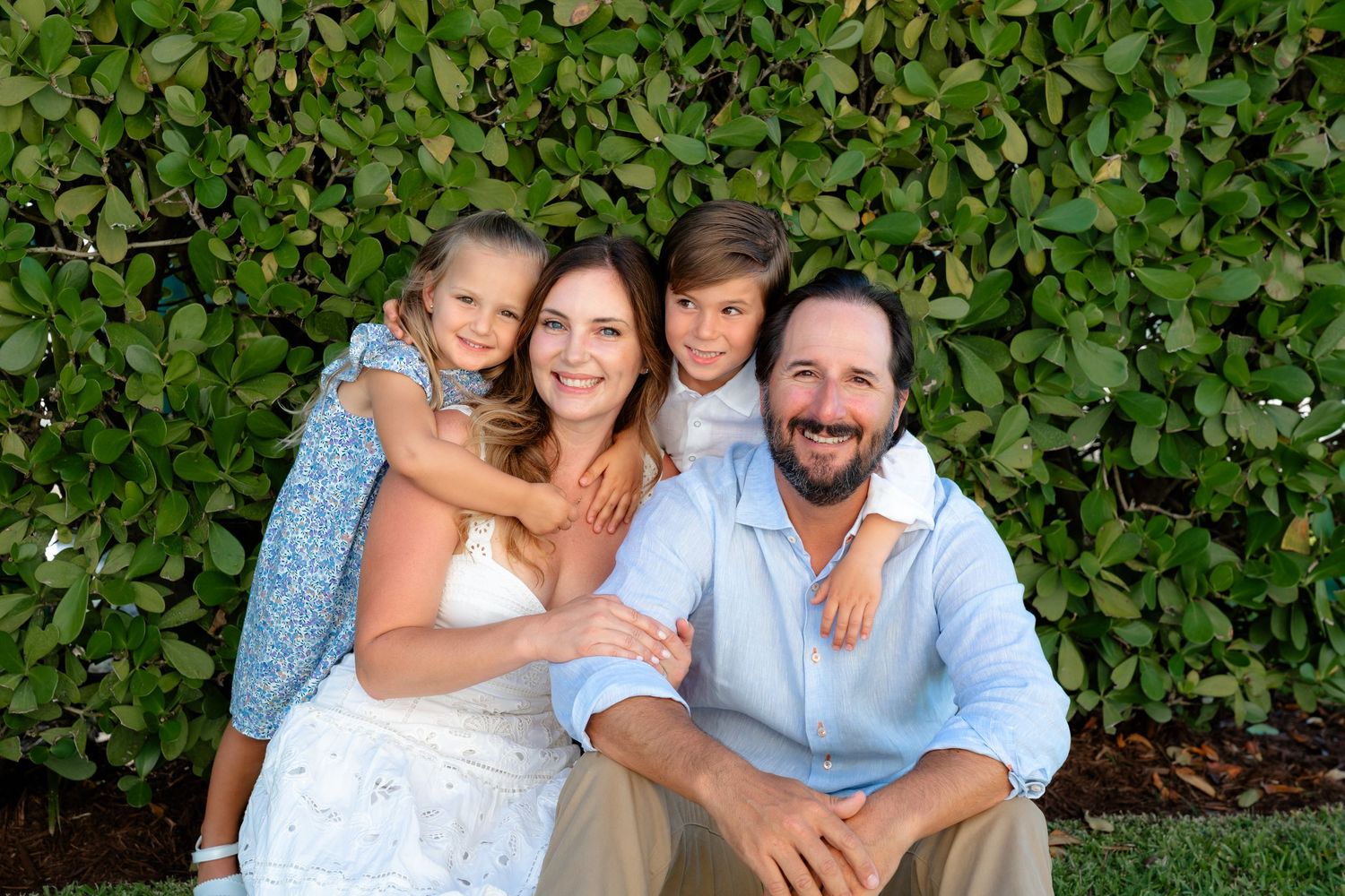 Happy family of four smiling together outdoors in front of lush green hedge on a sunny day.