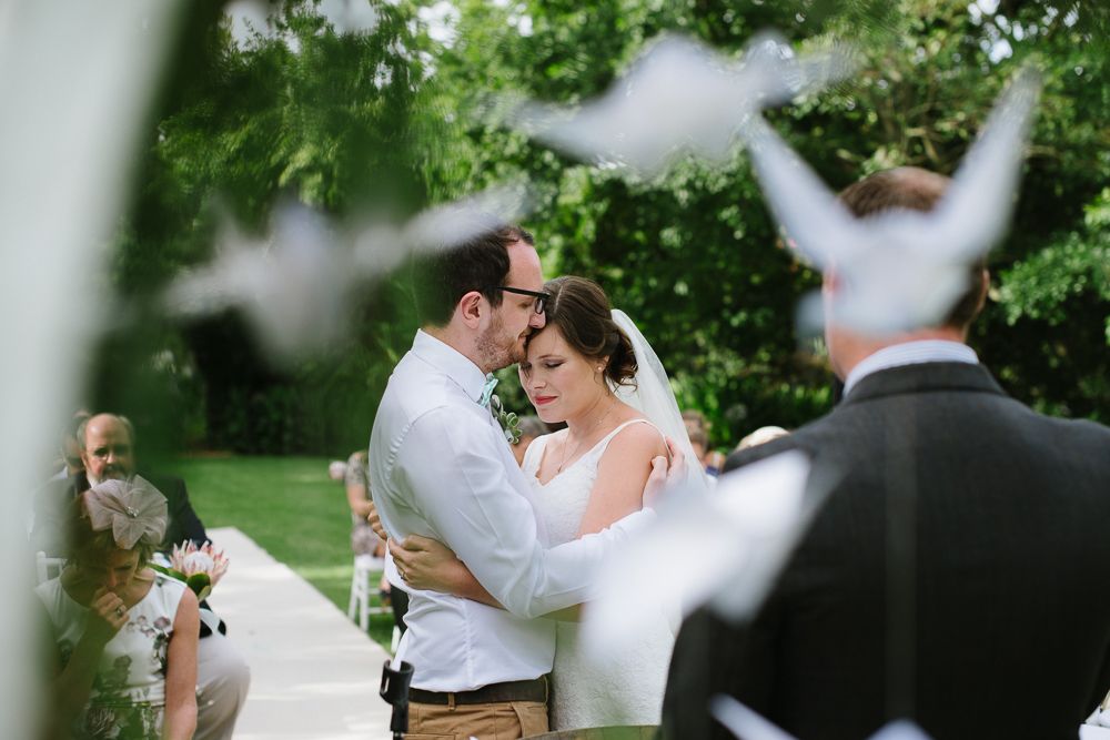 Bride and groom share an intimate embrace during outdoor wedding ceremony with white doves flying in the foreground.