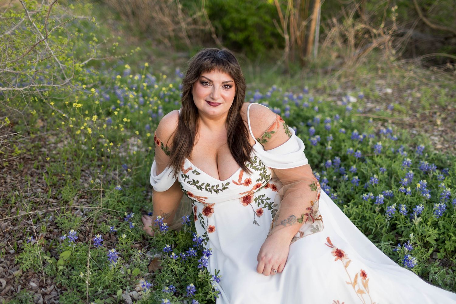 Woman in floral white off-shoulder dress sitting among bluebonnets and wildflowers in a sunlit garden setting.