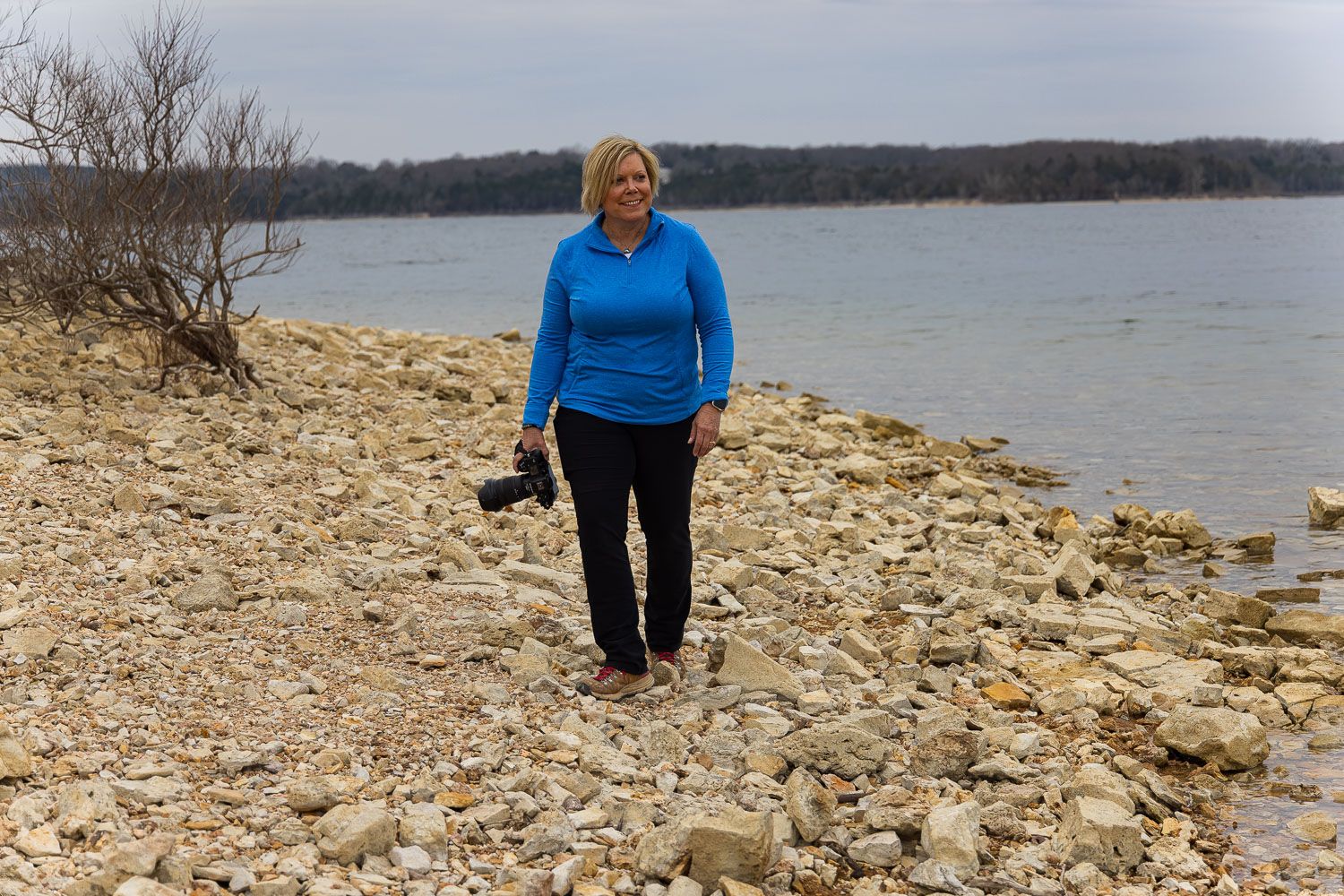 Woman in blue jacket walking on rocky shoreline near calm lake with bare trees in background.