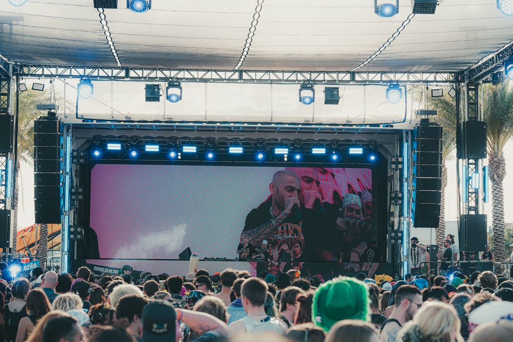 Large outdoor music festival crowd faces a bright stage with a giant screen displaying a performer.