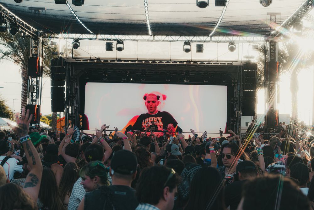 DJ performing on stage at outdoor music festival with large crowd and vibrant LED screen backdrop.
