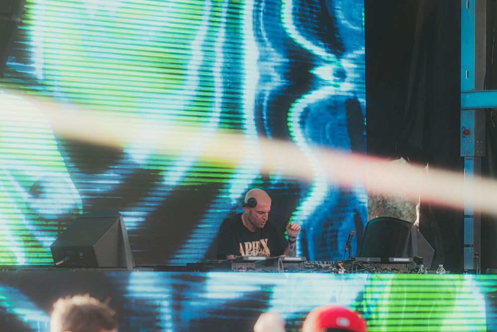 DJ performing on stage with vibrant green and blue psychedelic visuals backdrop at outdoor music festival.