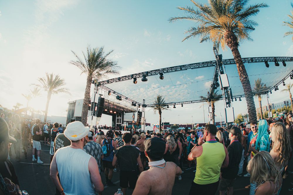 Crowd enjoys outdoor music festival near palm trees with stage lighting at sunset.