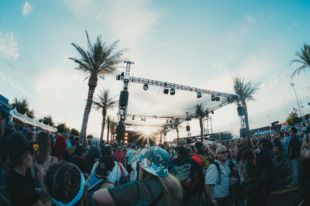 Festival crowd gathers at outdoor stage surrounded by palm trees during golden hour sunset.