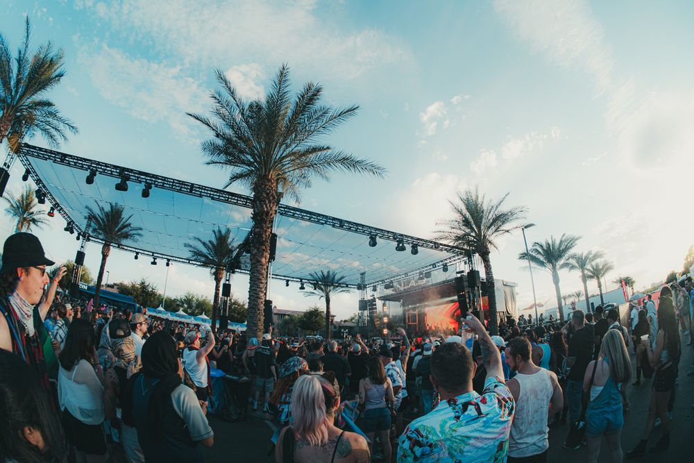 Outdoor music festival crowd with palm trees and a lit stage at sunset in a warm, vibrant atmosphere.