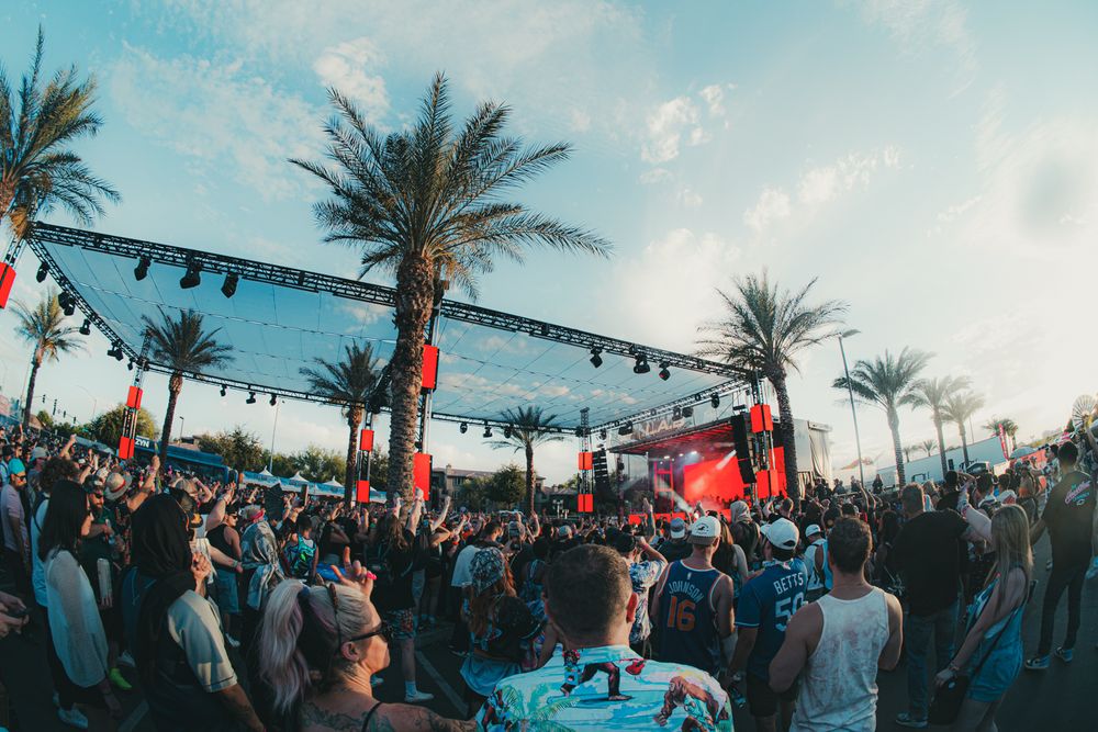 Outdoor music festival crowd with palm trees and colorful stage lighting at dusk.