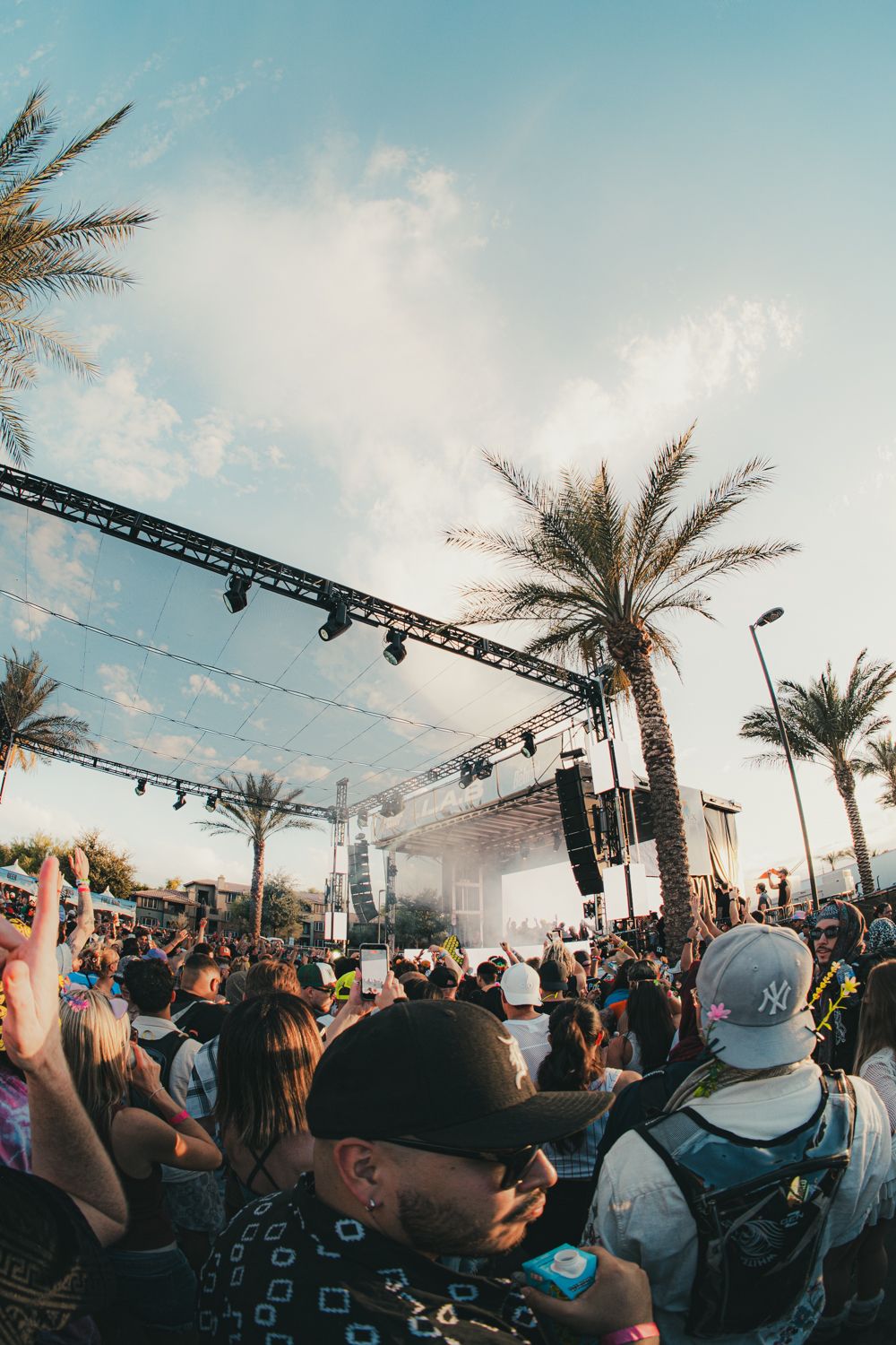 Crowd enjoys outdoor music festival stage with palm trees and dramatic sky in warm sunny weather.