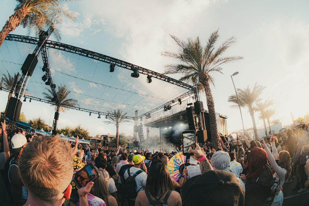 Outdoor music festival crowd cheering before a stage with palm trees and dramatic sky at sunset.