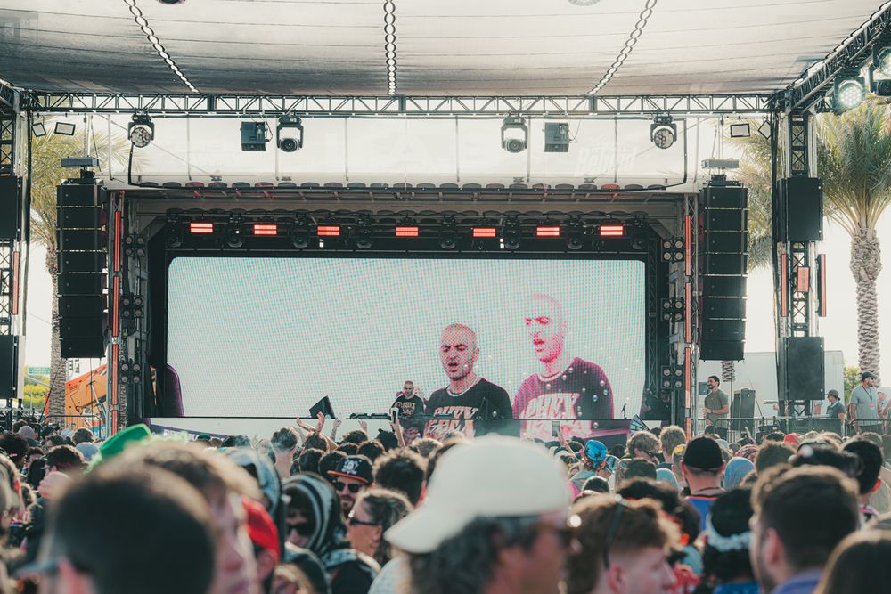 Outdoor music festival crowd watches two DJs perform on a large stage with bright LED screen backdrop.